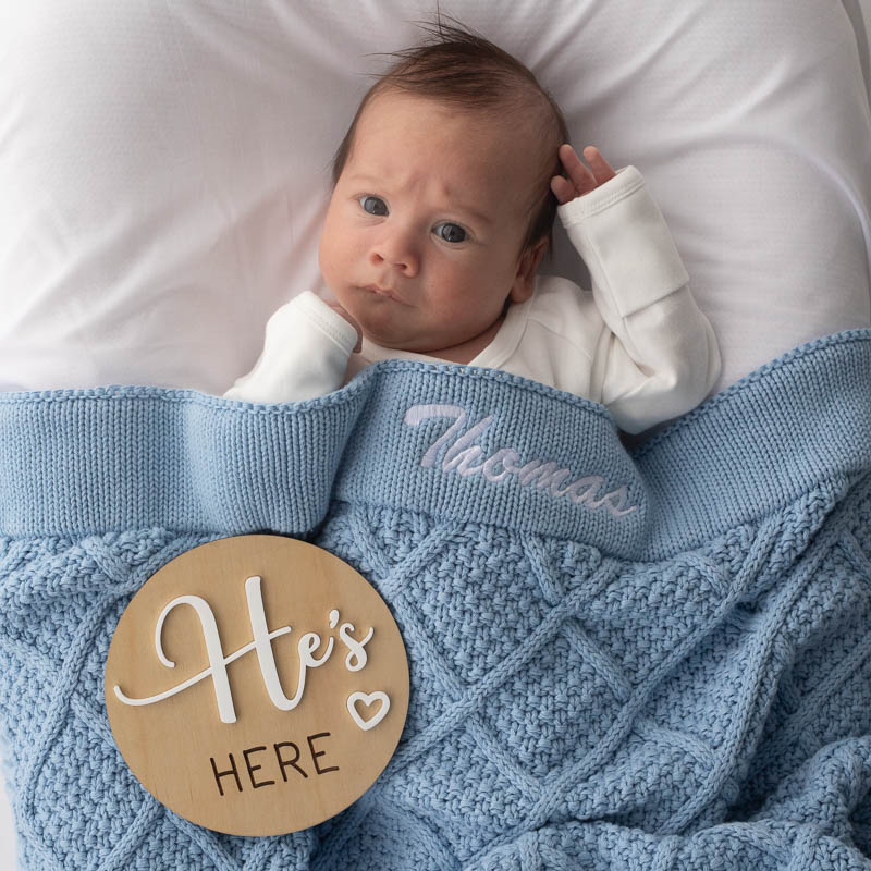Baby wrapped in personalised blue diamond blanket with “He’s Here” wooden announcement disc.