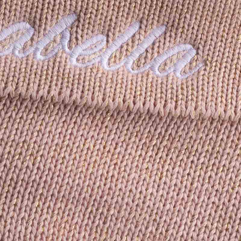 Close-up of pink knit blanket with subtle gold thread and name embroidered with white thread.