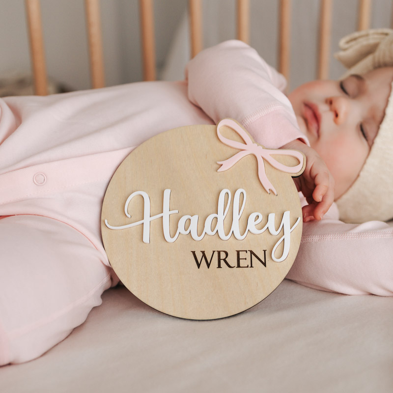 Sleeping baby in pink holds wooden birth disc with acrylic bow, reading “Hadley Wren.”