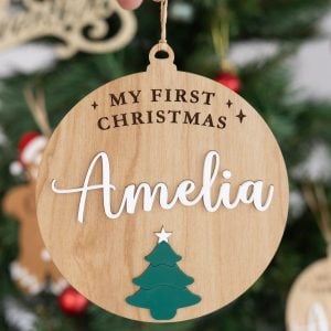 Close-up of personalised wooden Christmas Disc engraved with My First Christmas and Amelia in white with green tree detail