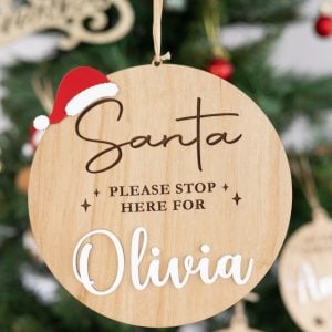 Close-up of personalised wooden Christmas bauble engraved Santa Please Stop Here For Olivia in white with red Santa hat detail