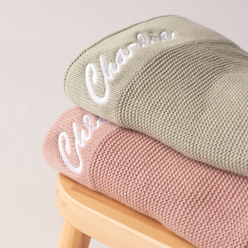Blush pink and sage green personalised knitted baby blankets stacked on wooden stool.