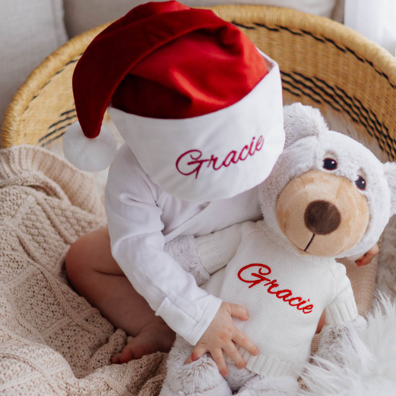 Baby wearing personalised Christmas hat cuddling matching teddy.