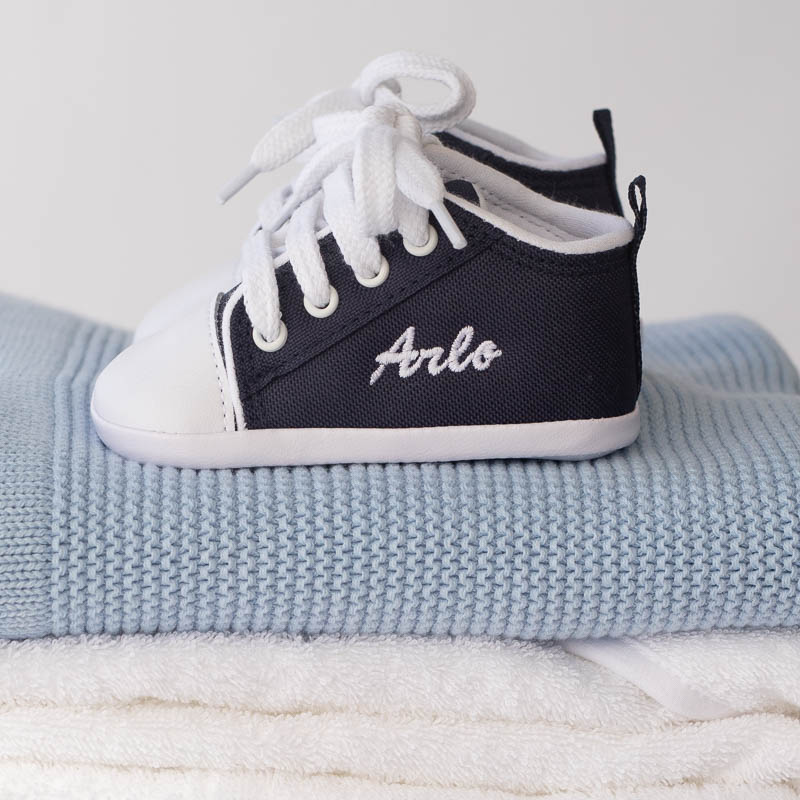Navy blue personalised baby shoes with “Arlo” embroidered, placed on a blue knitted blanket.