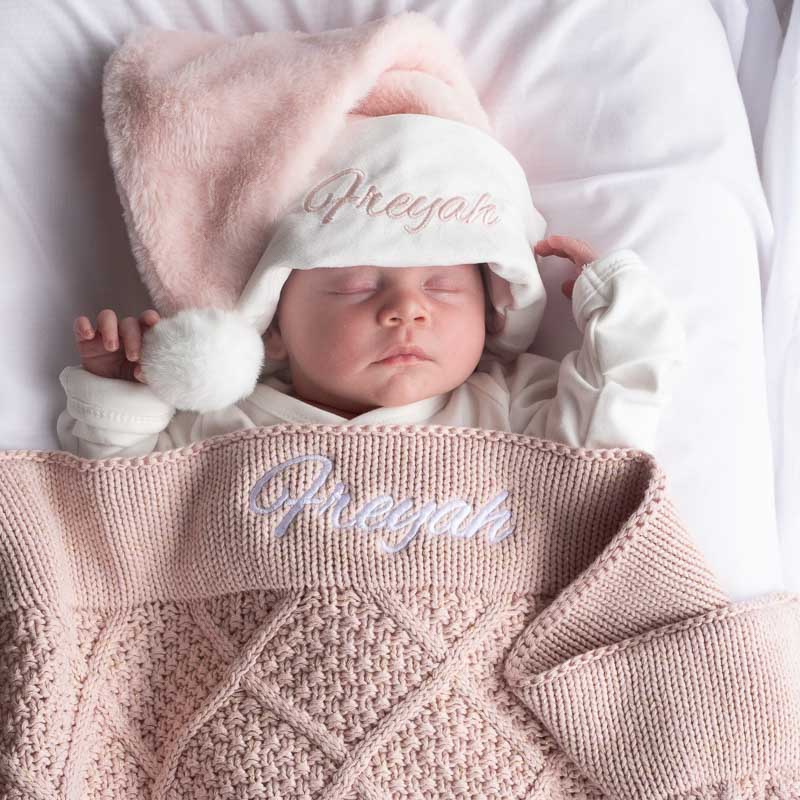 Baby wearing pink Christmas hat and blush pink diamond knitted blanket, sleeping peacefully on a soft surface.