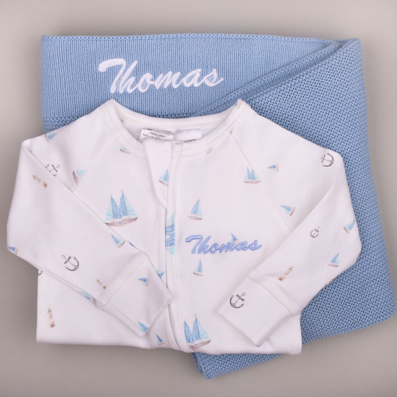 Blue Baby Blanket Nautical Onesie White nautical-print baby onesie with name ‘Thomas,’ displayed on a blue knitted blanket with matching embroidery.