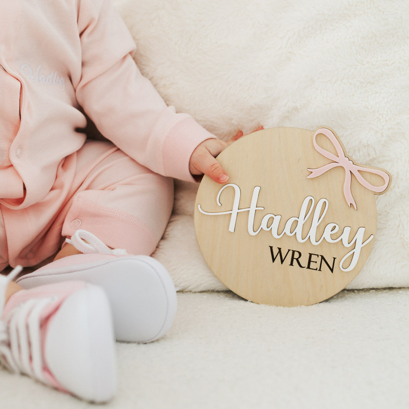 Baby in pink holding wooden bow name disc.