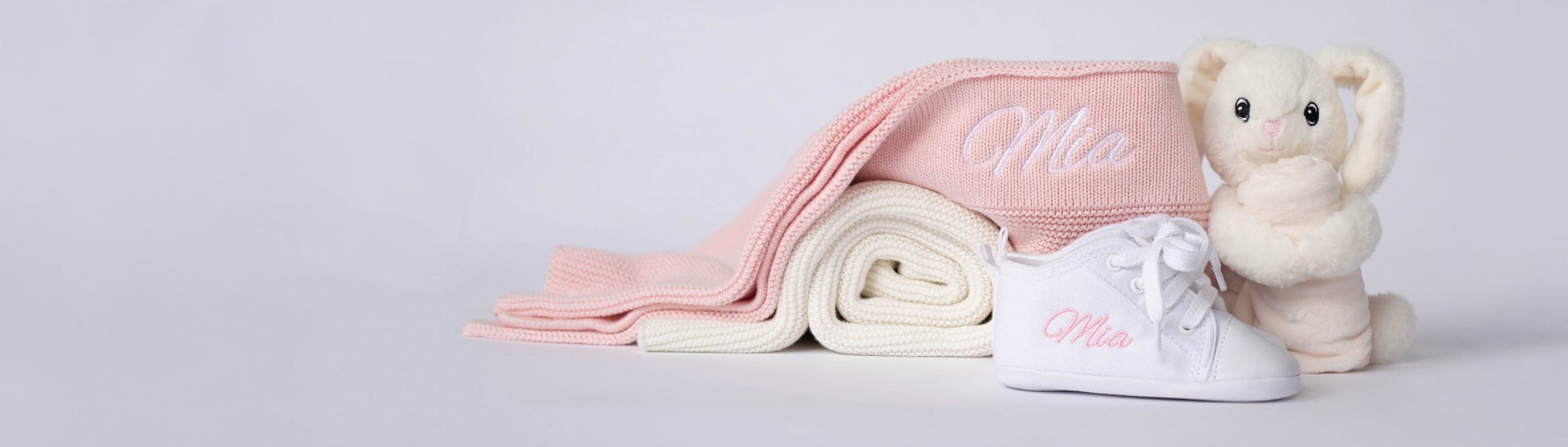 A white knitted baby blanket, bunny comforter & white shoes all embroidered with the name Mia.