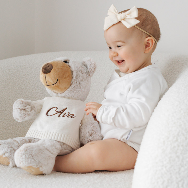Smiling baby in white romper with bow sitting beside personalised teddy bear with Ava jumper on white chair.