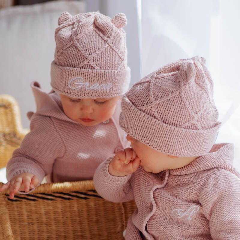 Two babies wearing matching blush pink personalised beanies and hooded cardigans in a wicker basket.