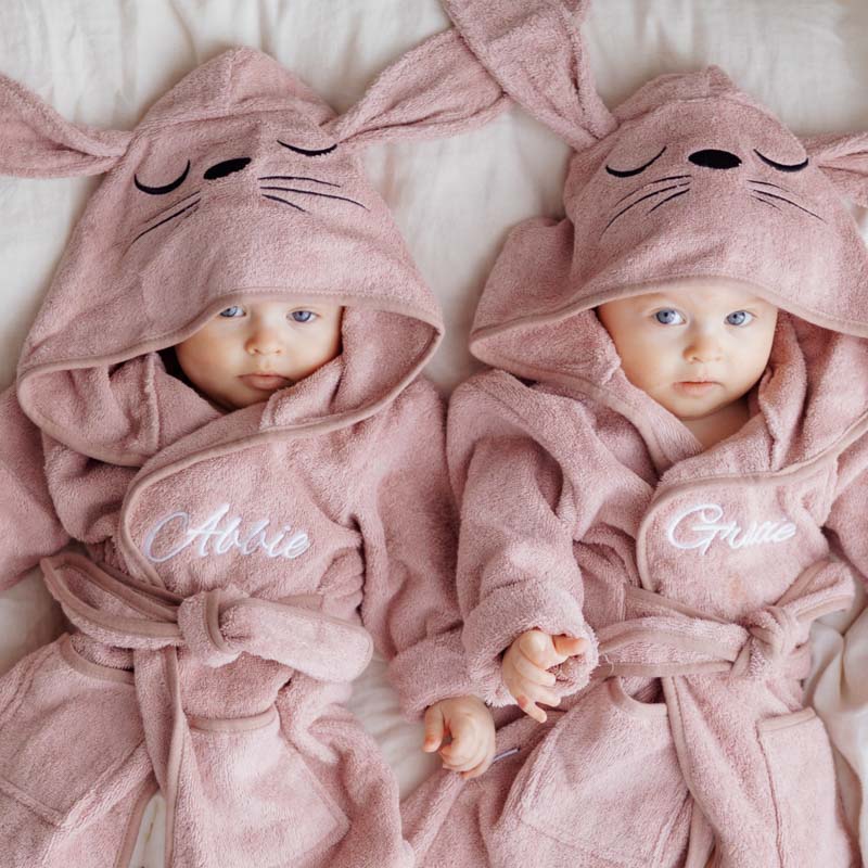 Two babies in pink personalised bunny hooded robes lying together on a soft bed.