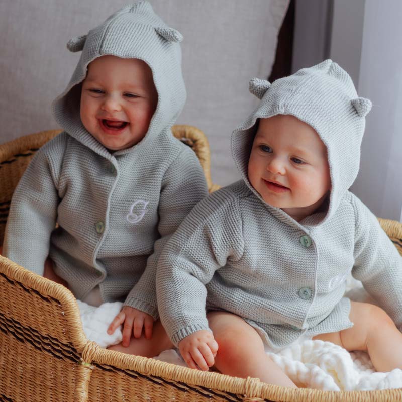 Twin babies in sage hooded personalised cardigans sitting in a wicker basket, smiling happily.