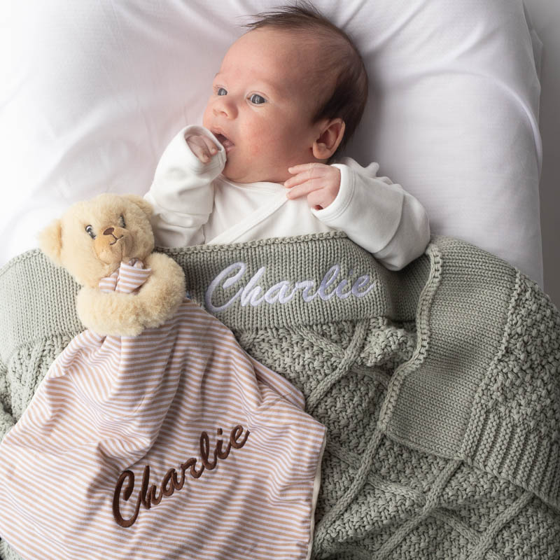 Newborn lying under personalised sage knitted blanket cuddling a striped bear comforter.