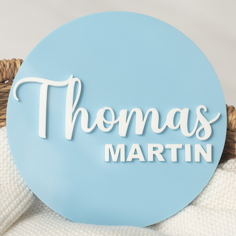 Blue acrylic baby name announcement disc with raised white lettering.