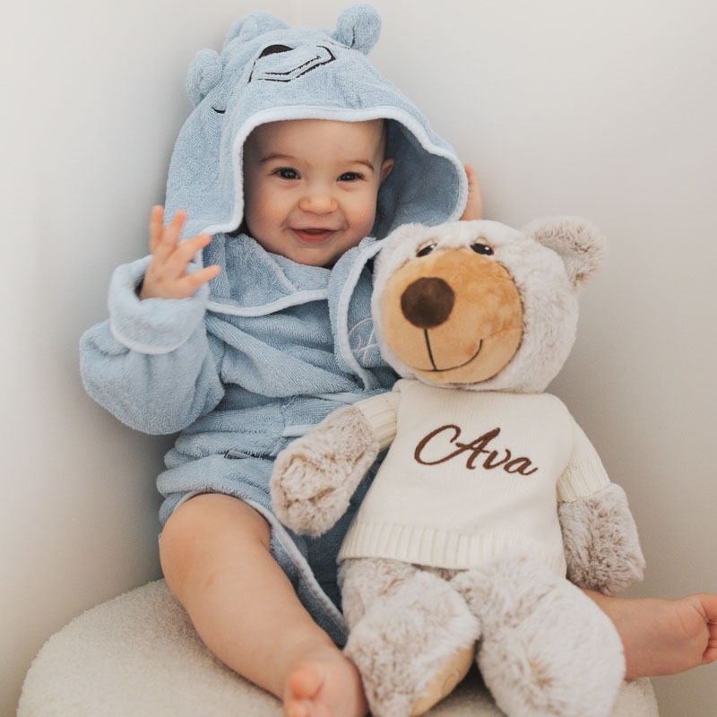 Baby in blue bear hooded robe holding personalised teddy bear.