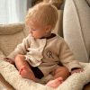 Beige Baby Cardigan Sitting Initial R Baby wearing monogram 'R' knit cardigan romper in beige, seated in cosy nursery chair.