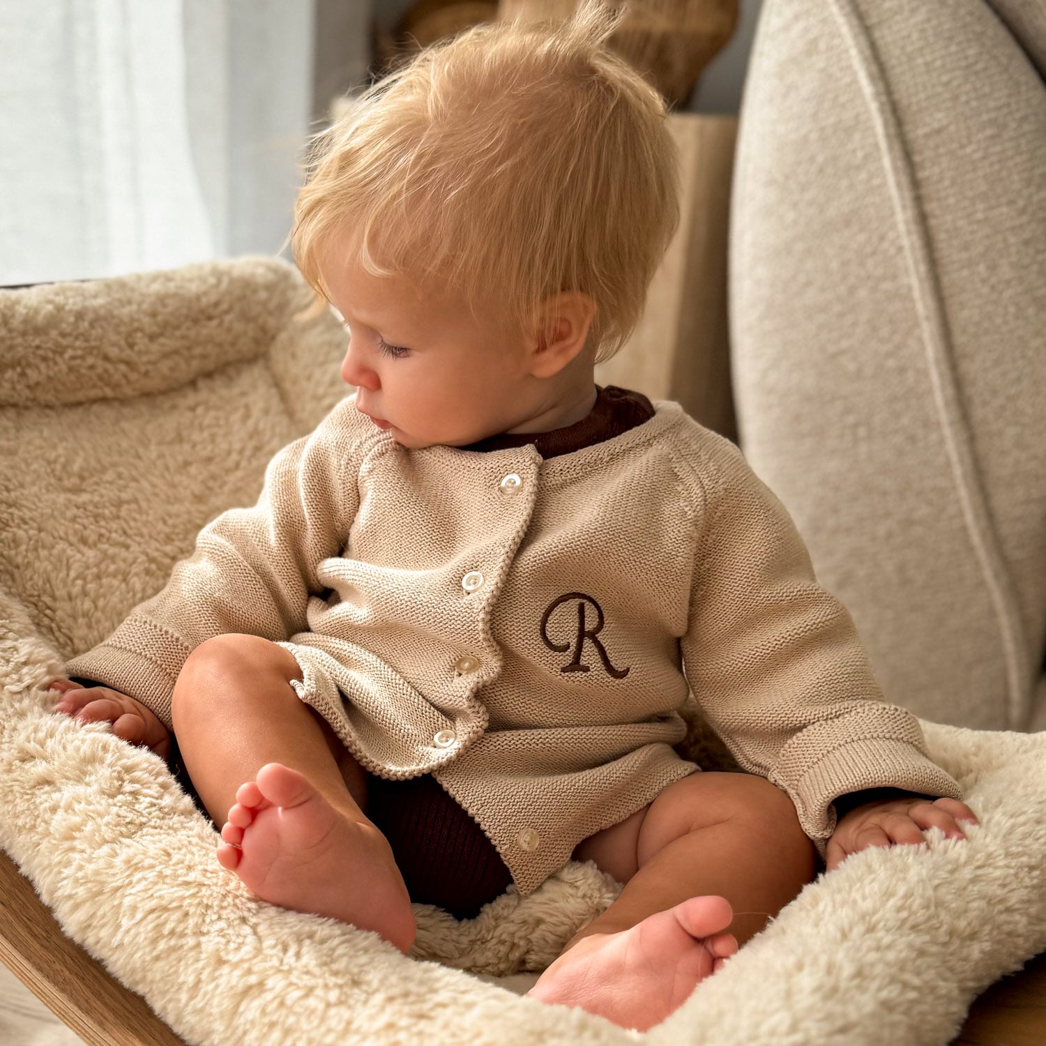 Baby wearing monogram 'R' knit cardigan romper in beige, seated in cosy nursery chair.