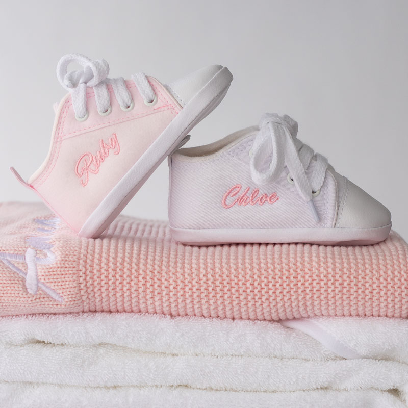 Personalised pink baby shoes embroidered with names, displayed on folded blankets.