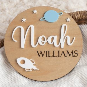 Round wooden name plaque reading “Noah Williams,” featuring stars, a blue planet, and a white rocket motif.