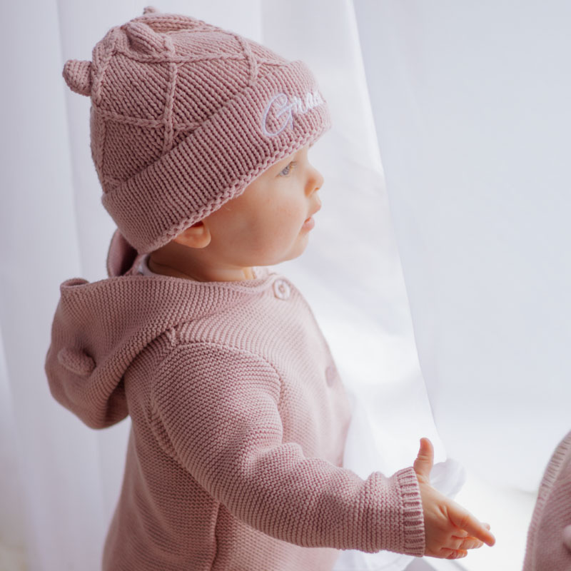 Baby wearing blush pink knitted beanie and hooded cardigan.