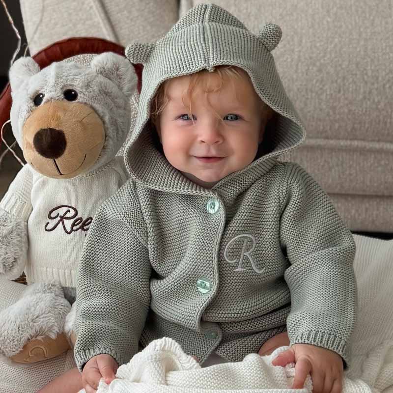 Baby in sage hooded cardigan sitting beside teddy in personalised jumper.