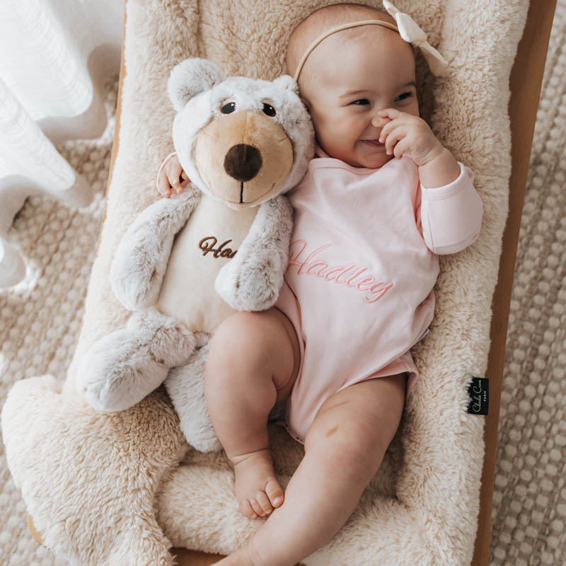 Smiling baby lying on a cushion holding a personalised teddy bear.