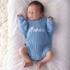 Pesonalised Blue Knitted Long Sleeve Baby Boy Romoer. Oscar Sleeping newborn wearing a blue knitted long sleeve baby romper with white embroidered script name on white bedding.