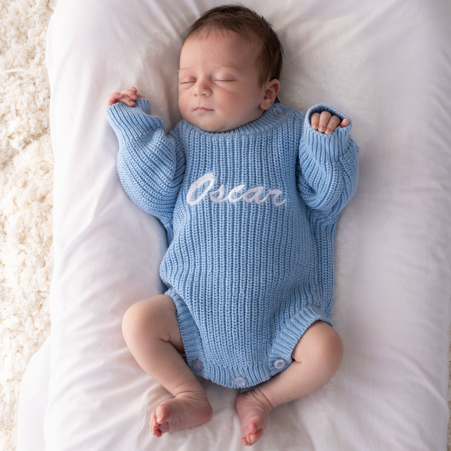 Sleeping newborn wearing a blue knitted long sleeve baby romper with white embroidered script name on white bedding.