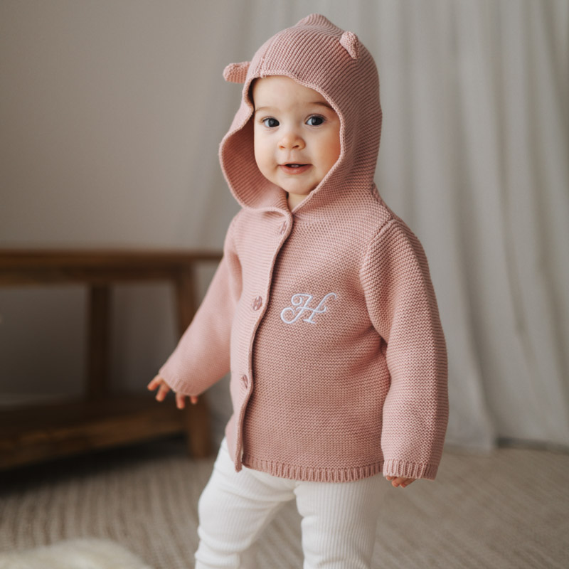 Baby wearing blush pink hooded knitted cardigan with initial.