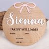 Round wood birth announcement disc with raised white acrylic name, pink acrylic bow and engraved full name and birth detail fields on wicker basket.