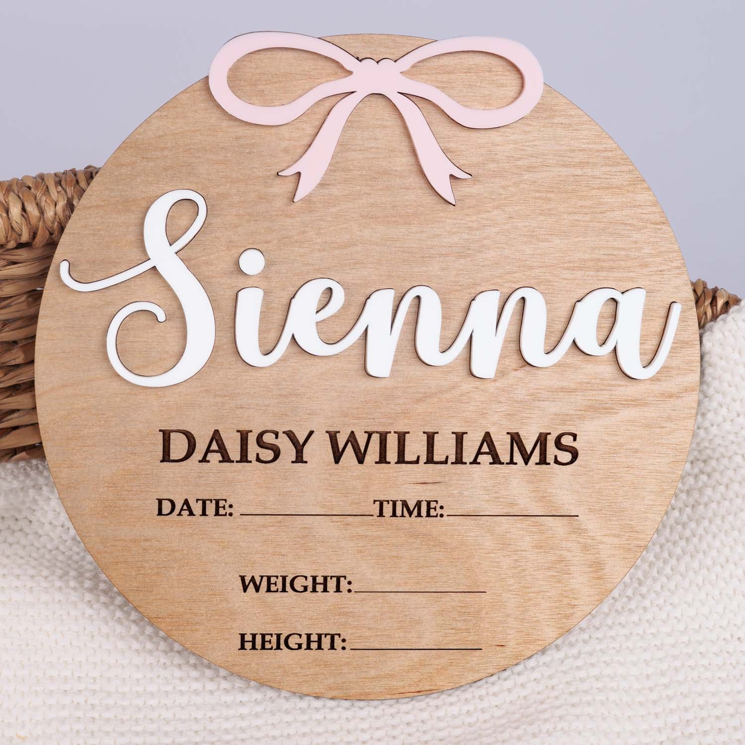 Round wood birth announcement disc with raised white acrylic name, pink acrylic bow and engraved full name and birth detail fields on wicker basket.