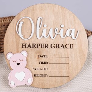 Round wood birth announcement disc with raised white acrylic name, pink acrylic bear and engraved full name and birth detail fields on wicker basket.