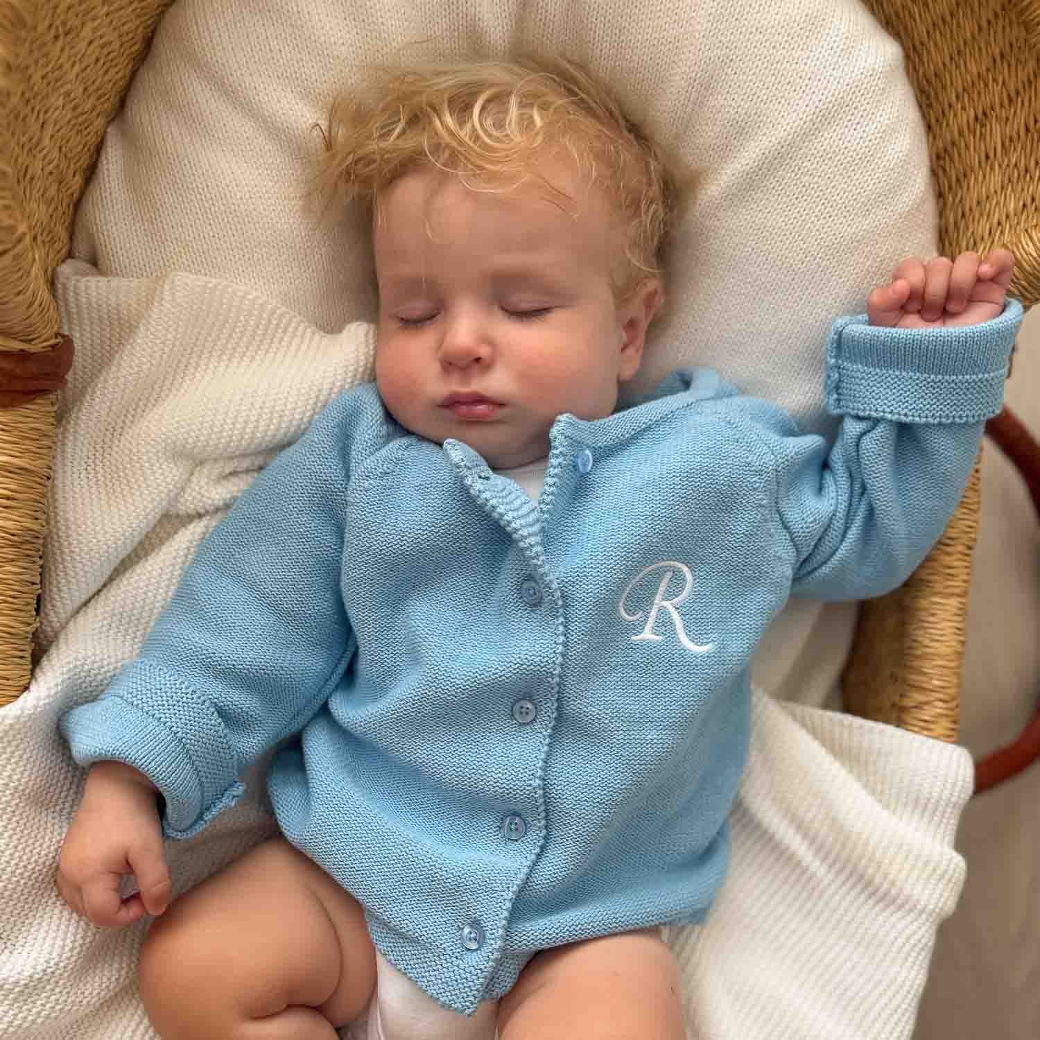 Sleeping baby wearing a personalised blue knitted cardigan with an embroidered initial, lying in a bassinet.