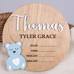 Round wood birth announcement disc with raised white acrylic name, blue acrylic bear and engraved full name and birth detail fields on wicker basket.