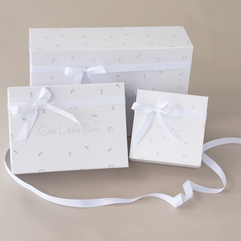 Three One Little Day gift boxes in varying sizes tied with white satin ribbon bows on a beige background.