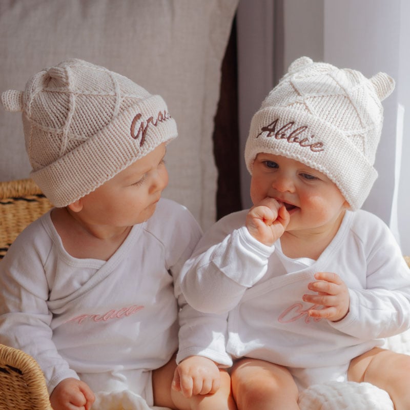 Two babies wearing personalised knitted diamond beanies with embroidered names in cream and beige.
