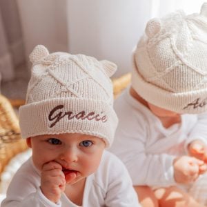 Two babies wearing cream diamond knitted beanies with ears, embroidered with the names Gracie and Archie.