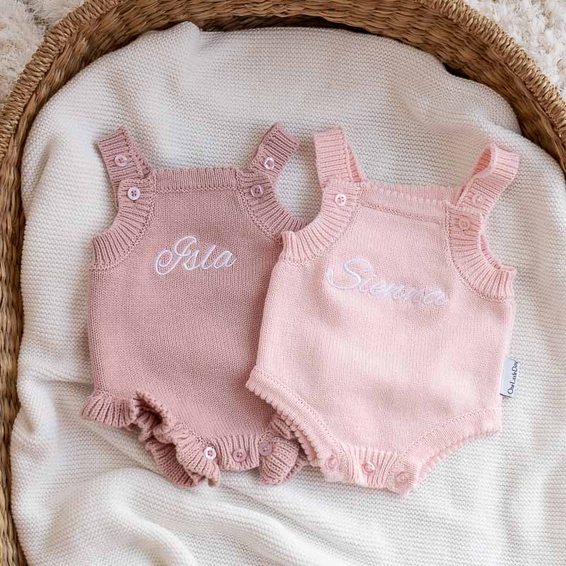 Two personalised pink frill knit baby girl rompers with embroidered names in a wicker basket.