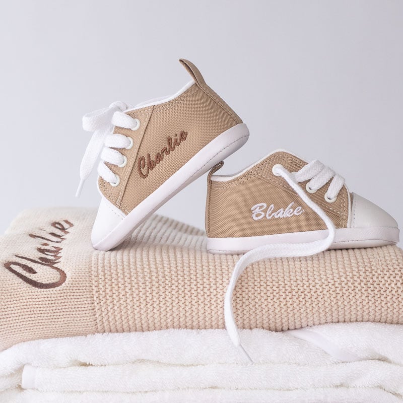 Personalised sand canvas baby sneakers with embroidered names stacked on a beige knit baby blanket.