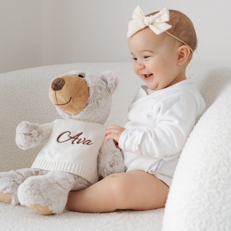 Baby smiling beside personalised teddy bear with knitted jumper.