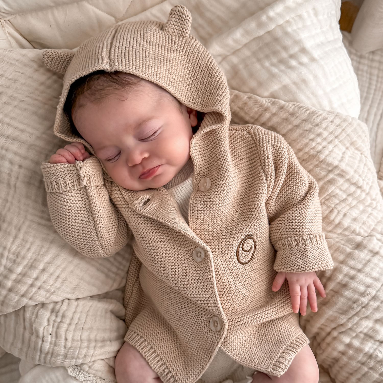 A baby wearing a Beige Hooded Knitted Baby Cardigan monogrammed with the letter O.