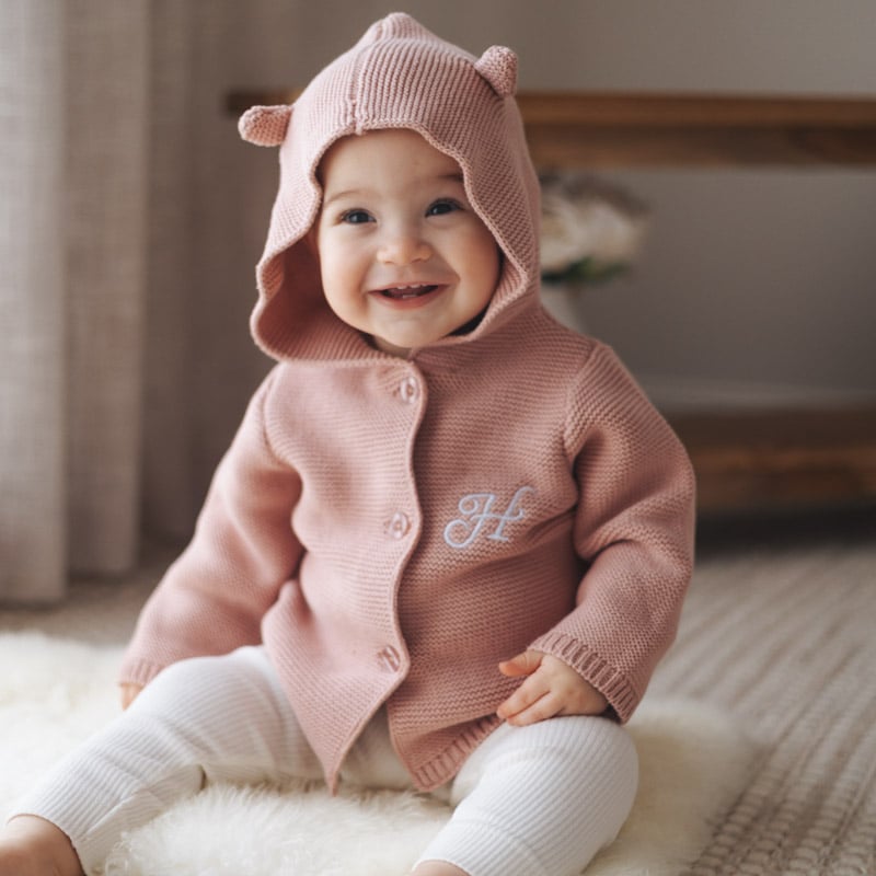 Smiling baby wearing blush pink hooded knitted cardigan.