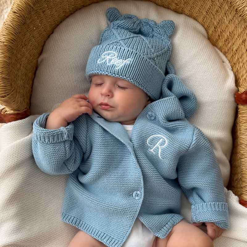 Sleeping newborn baby boy wearing a personalised blue knitted cardigan embroidered with the initial R and a matching blue diamond beanie embroidered with the name Reef.