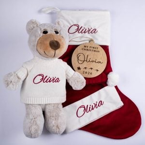 Four piece personalised Christmas baby gift set including grey plush teddy bear in white knitted jumper, burgundy velvet stocking, Santa hat with white cuff and wooden My First Christmas disc all with burgundy embroidered script name.