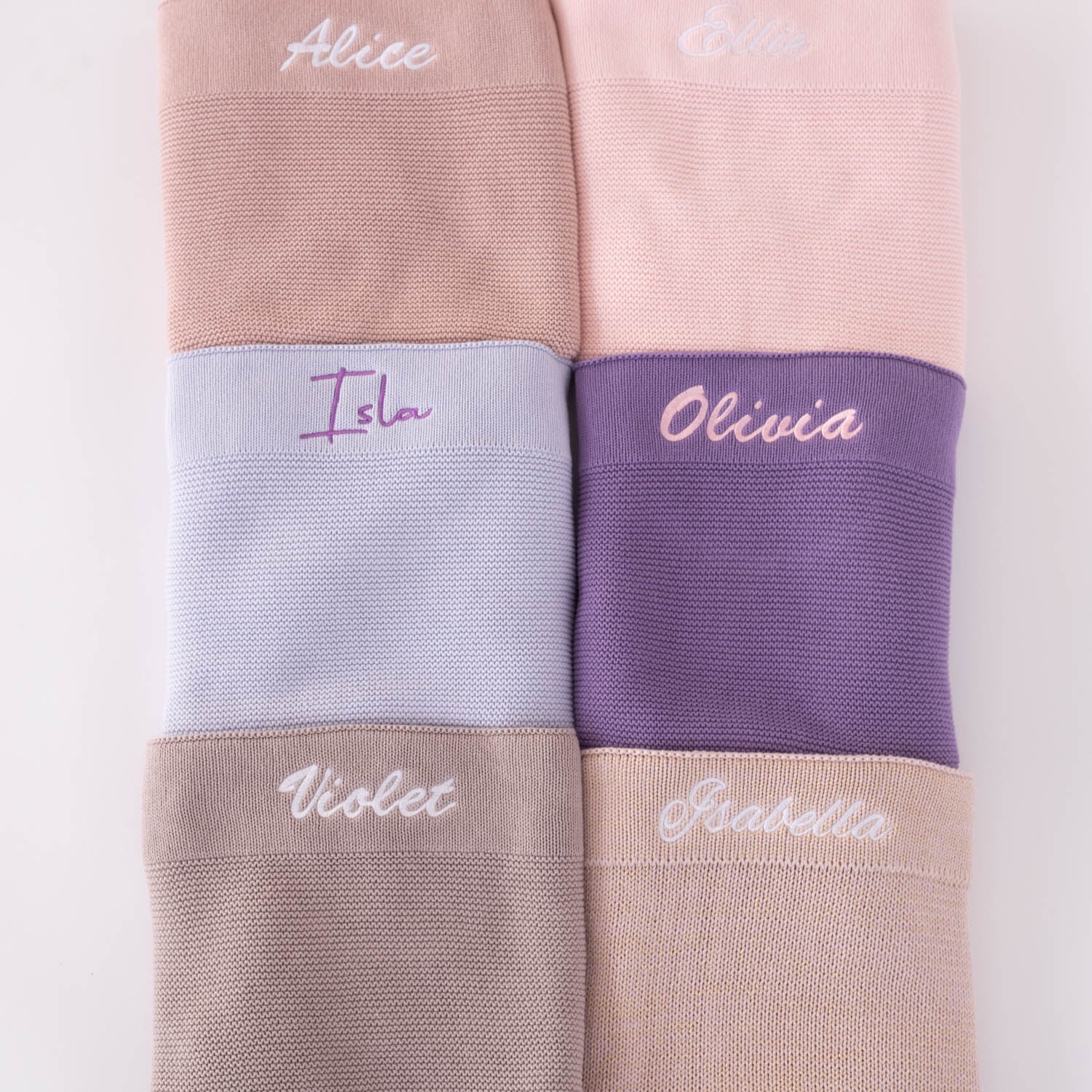 Six personalised knitted baby blankets in pink, lavender, dusty blue, deep purple, taupe and beige tones with embroidered script names in contrasting thread colours.