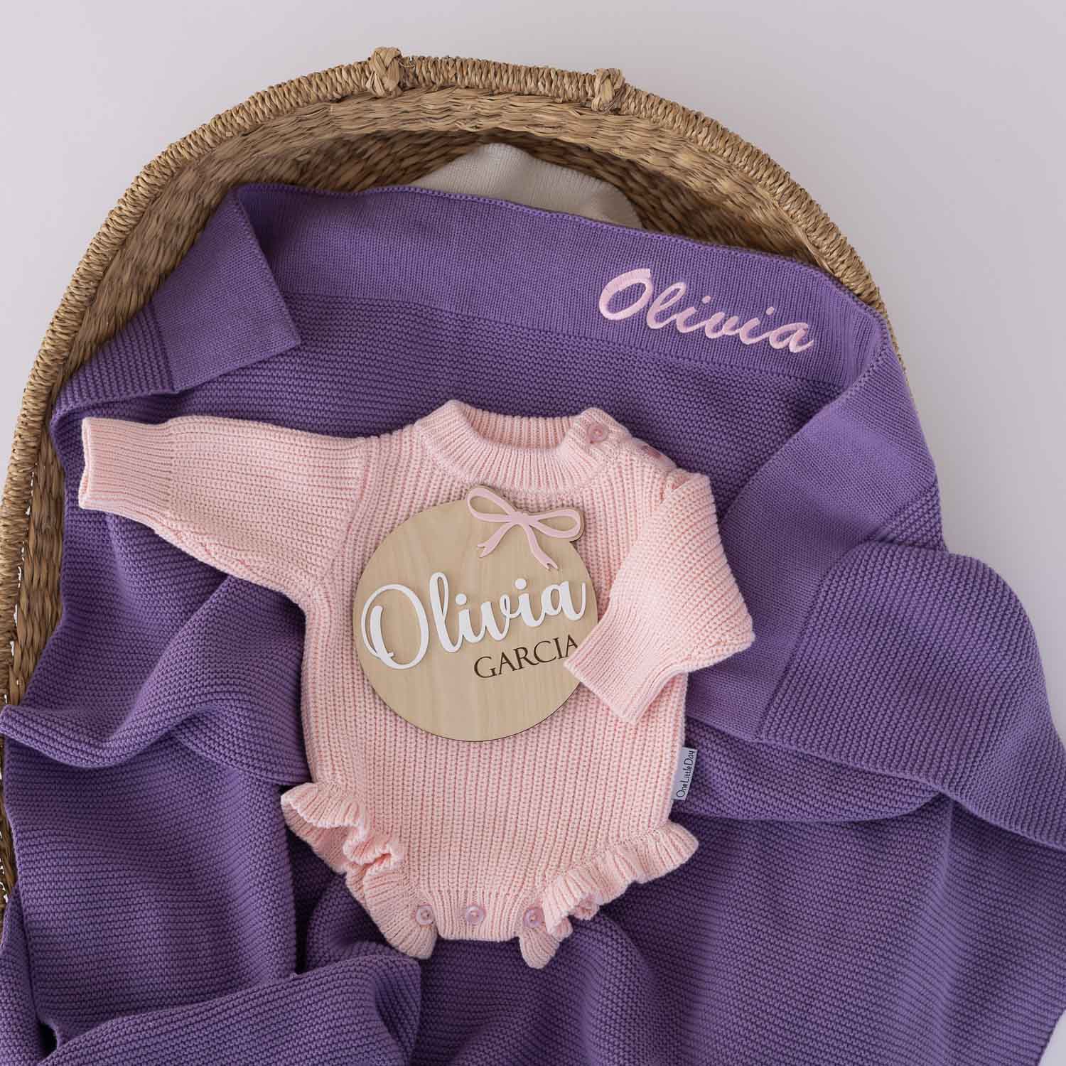 Personalised purple knitted baby blanket embroidered with the name Olivia displayed in a wicker bassinet with a blush pink knitted long sleeve romper and timber birth announcement disc personalised with Olivia Garcia.