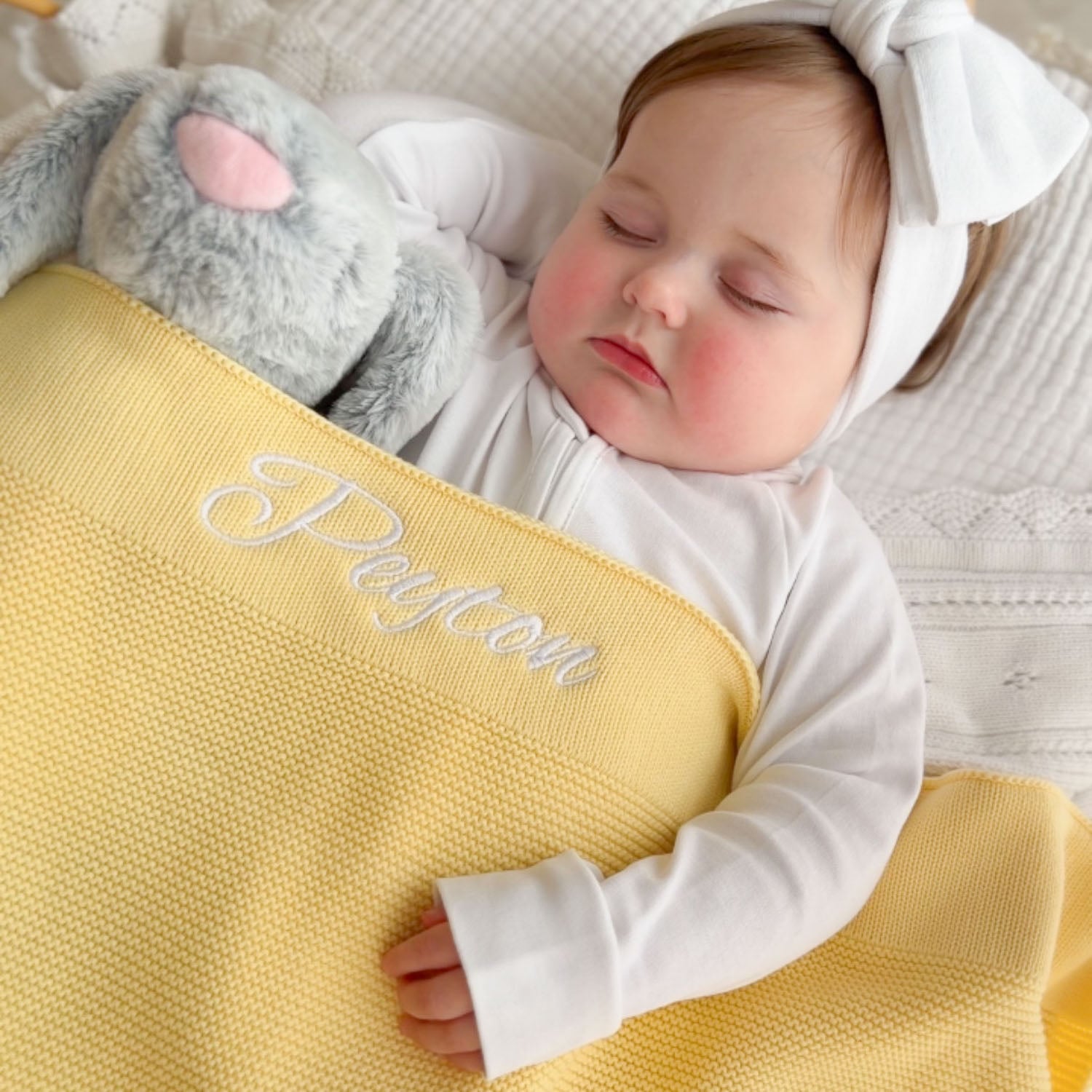Sleeping baby girl holding a personalised butter yellow knitted blanket embroidered with the name Peyton in white script alongside a personalised grey bunny in a white jumper.