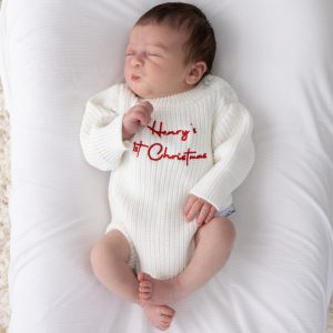 Sleeping newborn wearing a white knitted long sleeve baby romper with red embroidered personalised 1st Christmas script text on white bedding.