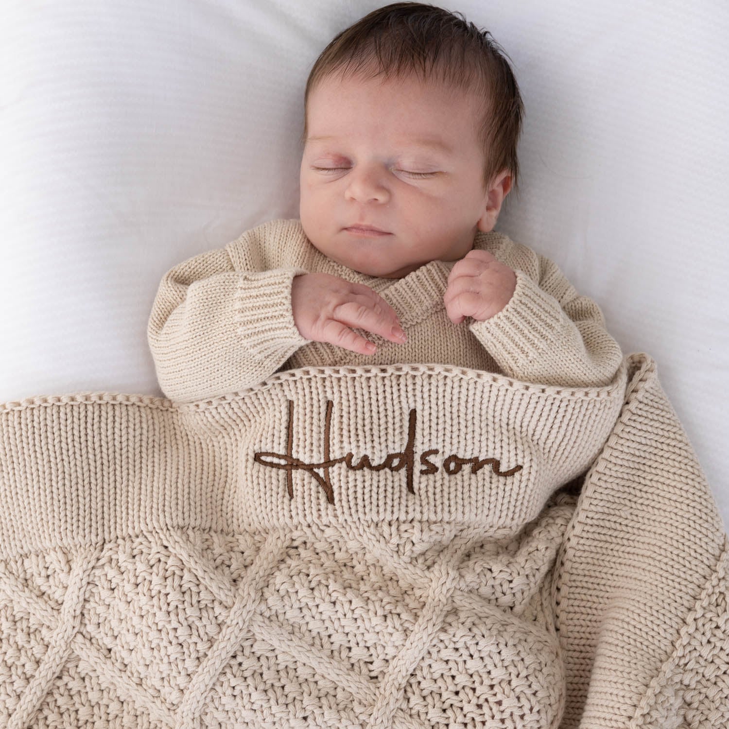 Sleeping newborn baby wrapped in a personalised beige diamond knitted baby blanket embroidered with the name Hudson in brown script.