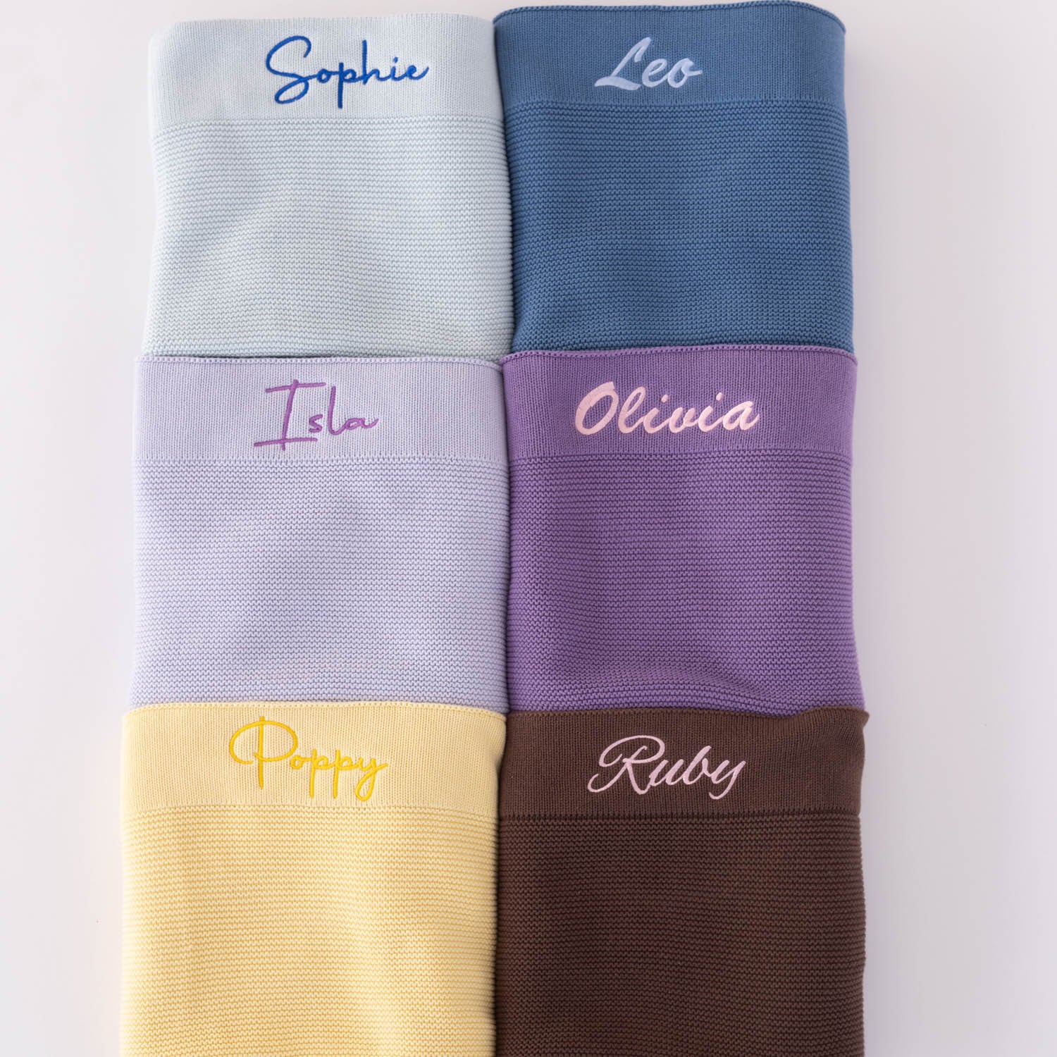 Six personalised knitted baby blankets in Pastel blue, dark blue, lavender, purple, Butter yellow and chocolate brown showing colour and embroidery font style comparison.